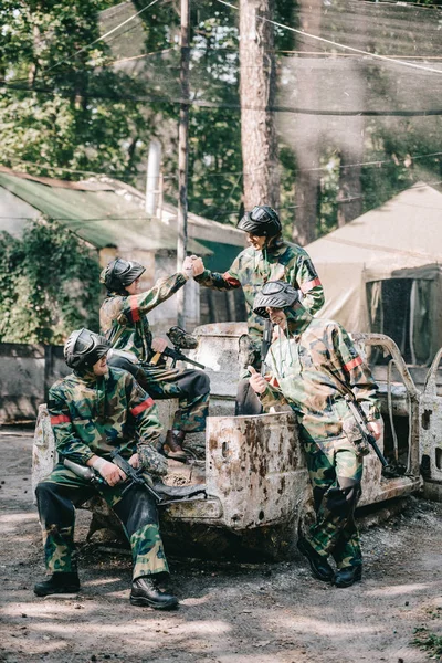 Equipo de paintball en camuflaje y máscaras de gafas celebrando la victoria y estrechando la mano en el coche roto al aire libre - foto de stock