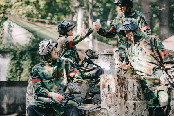 Happy paintball team in camouflage celebrating victory and shaking hands on broken car outdoors — Stock Photo