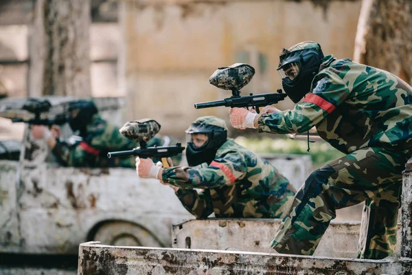 Paintball players in camouflage and protective masks aiming with marker guns from broken car outdoors — Stock Photo
