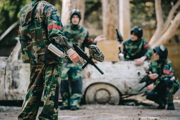 Imagen recortada del equipo de paintball en uniforme de camuflaje con pistolas marcadoras descansando cerca de coche roto al aire libre - foto de stock