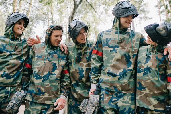 Sonrientes jugadores de paintball en camuflaje con pistolas marcadoras hablando y abrazándose al aire libre - foto de stock