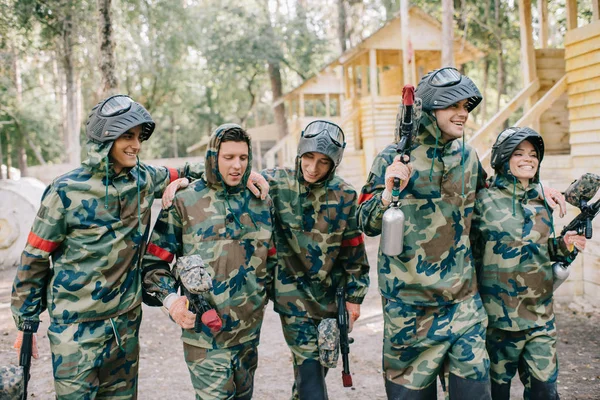 Riéndose jugadores de paintball en camuflaje con pistolas marcadoras abrazándose al aire libre - foto de stock