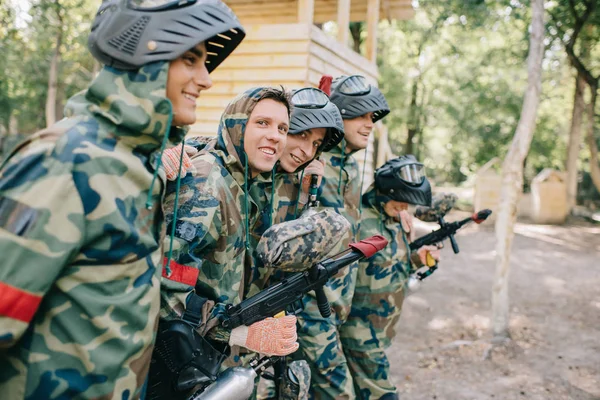 Selective focus of happy paintball players in camouflage with marker guns embracing each other outdoors — Stock Photo