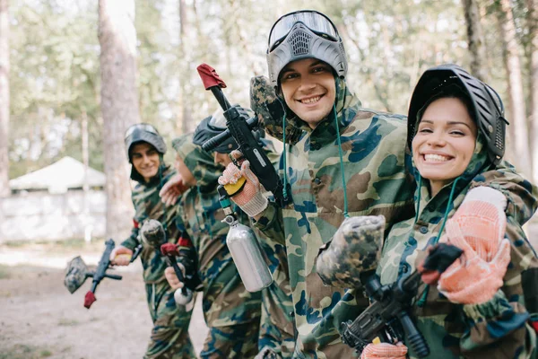 Sorridente jovem pintor masculino abraçando companheiro de equipe feminino em camuflagem com pistola de paintball ao ar livre — Fotografia de Stock