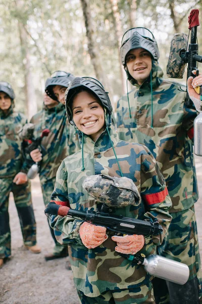 Hermosa mujer feliz paintballer en uniforme sosteniendo pistola de paintball mientras su equipo de pie detrás al aire libre - foto de stock