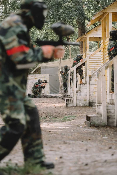 Selective focus of paintballer in camouflage and protective mask aiming by marker gun while his team standing behind outdoors — Stock Photo