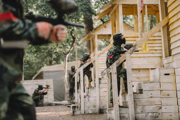 Equipo de paintball en uniforme y máscaras protectoras de pie en la escalera de torres de madera con pistolas de paintball al aire libre - foto de stock