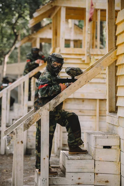 Female paintballer in camouflage and goggle mask with marker gun standing on staircase of wooden tower outdoors — Stock Photo