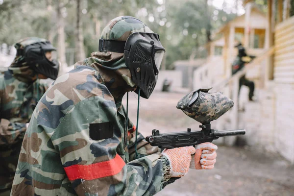 Vista lateral do pintor masculino e sua equipe em máscaras uniformes e protetoras visando por pistolas de paintball ao ar livre — Fotografia de Stock