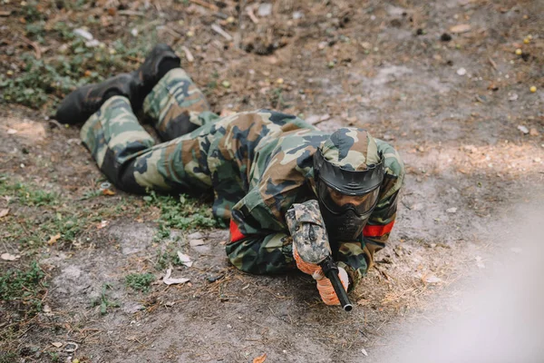 Hochwinkelaufnahme des männlichen Paintballspielers in Brillenmaske und Tarnung kriechend mit Paintballpistole im Freien — Stockfoto