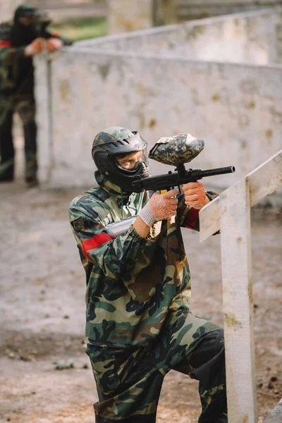 Side view of confident male paintball player in goggle mask and camouflage aiming by paintball gun outdoors — Stock Photo