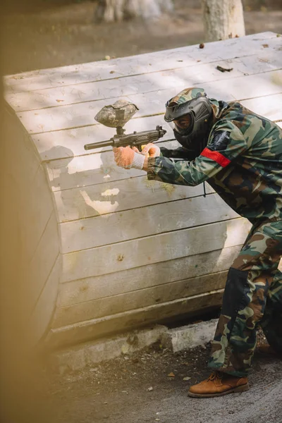 Vista lateral del jugador de paintball enfocado en máscara de gafas y camuflaje con el objetivo de pistola de paintball al aire libre - foto de stock