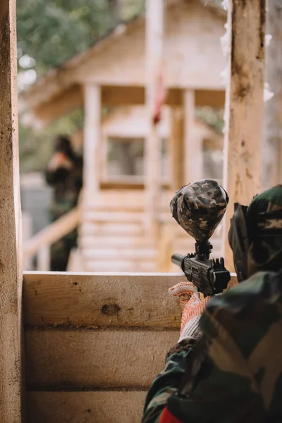 Vista parcial del jugador de paintball en uniforme de camuflaje apuntando con pistola de paintball desde torre de madera al aire libre - foto de stock