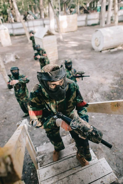 High angle view of male paintball player in goggle mask with marker gun on staircase while his team standing behind outdoors — Stock Photo