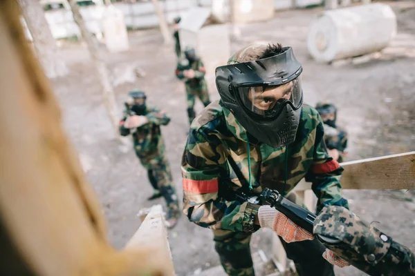 Jugador de paintball masculino en máscara de gafas y camuflaje con pistola marcadora en la escalera mientras su equipo de pie detrás al aire libre - foto de stock