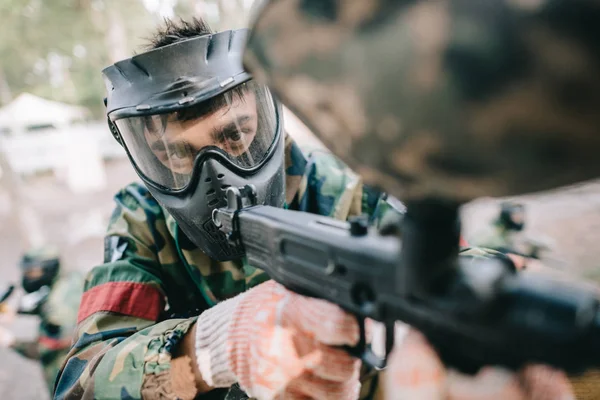 Selective focus of male paintball player in goggle mask and camouflage aiming by paintball gun outdoors — Stock Photo