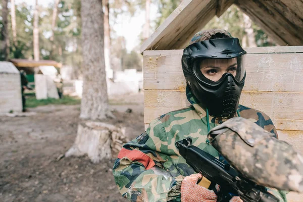 Serio verniciatore donna in maschera protettiva e uniforme mimetica in piedi con pistola paintball all'aperto — Foto stock