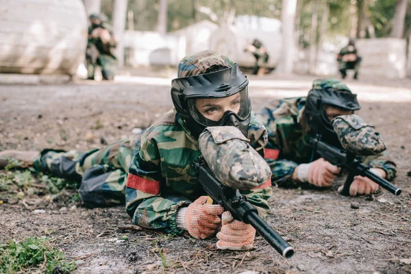 Serious female paintball player in goggle mask and camouflage with marker gun crawling on ground near teammate outdoors — Stock Photo