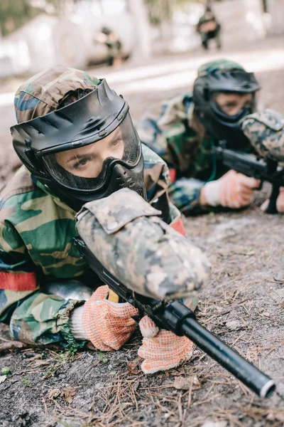 Female paintball player in goggle mask and camouflage with marker gun crawling on ground with teammate outdoors — Stock Photo
