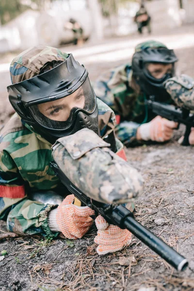 Mujer jugadora de paintball en máscara de gafas y camuflaje con pistola marcadora arrastrándose en el suelo con su compañero de equipo al aire libre - foto de stock