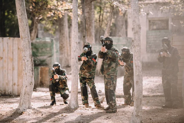 Selective focus of paintball team in uniform and protective masks aiming by paintball guns outdoors — Stock Photo