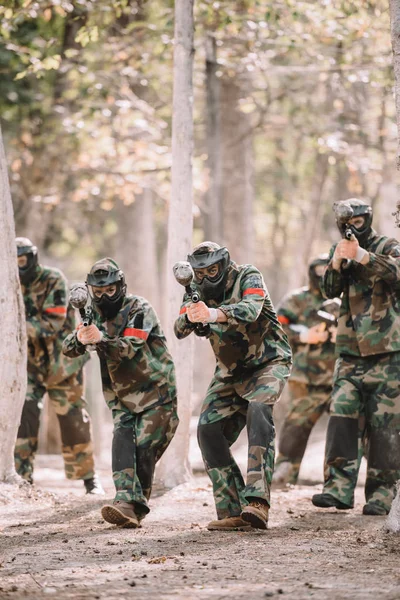 Команда пейнтболу в уніформі та захисних масках, що працюють та прицілюються пістолетами-пейнтболами на відкритому повітрі — стокове фото
