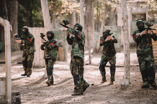 Команда з пейнтболу в уніформі та захисних масках, які стріляють пістолетами з пейнтболу на відкритому повітрі — стокове фото
