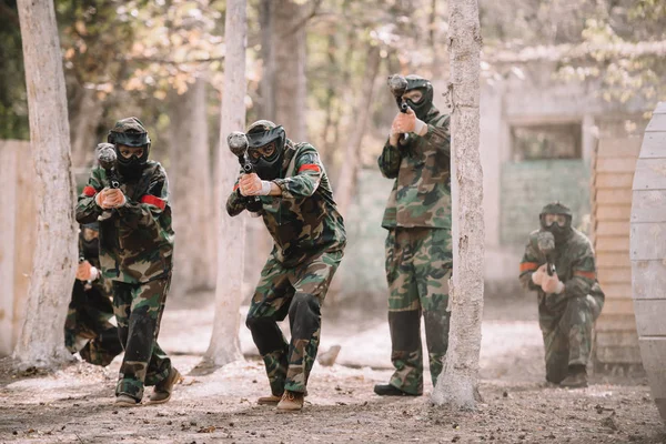 Paintball team in uniform and protective masks running and aiming by paintball guns outdoors — Stock Photo