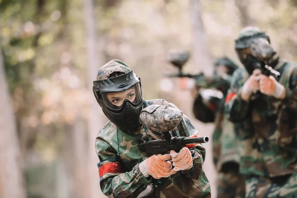 Female paintballer in goggle mask and camouflage shooting by marker gun while her team running behind outdoors — Stock Photo