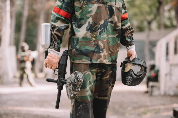 Cropped image of paintball player holding marker gun and goggle mask covered by paintball splash outdoors — Stock Photo