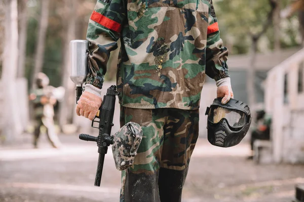 Imagen recortada del jugador de paintball sosteniendo pistola marcador y máscara de gafas cubiertas por salpicaduras de paintball al aire libre - foto de stock