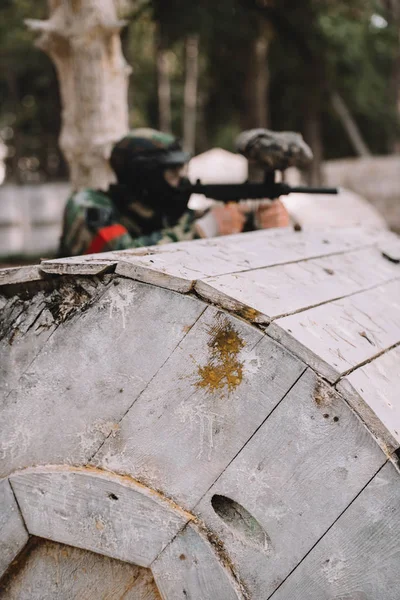 Selective focus of paintball splashes on wooden barrel and paintball player shooting behind outdoors — Stock Photo