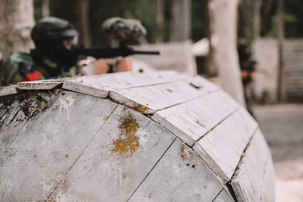 Selective focus of paintball splashes on wooden barrel and paintball player shooting behind outdoors — Stock Photo