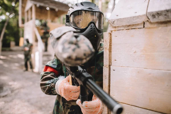 Selective focus of male paintballer in goggle mask covered by paintball splash aiming by marker gun outdoors — Stock Photo