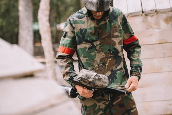 Male paintball player with marker gun looking at camouflage covered by paintball splash outdoors — Stock Photo