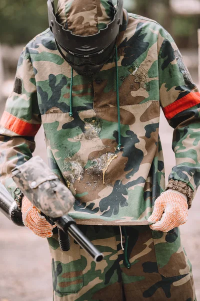 Paintball player with marker gun looking at camouflage covered by paintball splash outdoors — Stock Photo