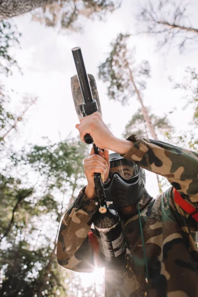 Low angle view of male paintball player in goggle mask and camouflage aiming by paintball gun outdoors — Stock Photo