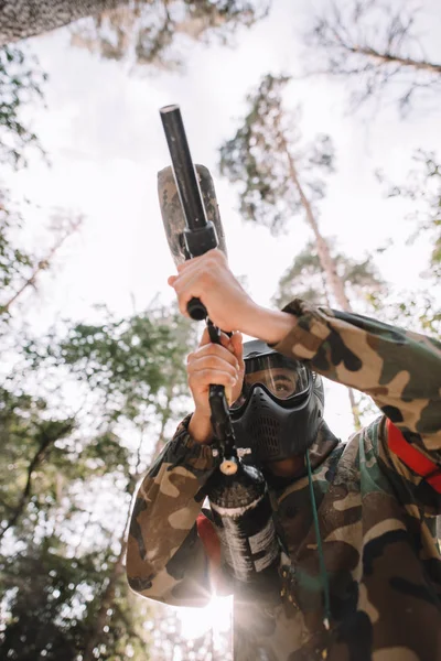 Vista de ángulo bajo del jugador de paintball masculino en máscara de gafas y camuflaje con el objetivo de pistola de paintball al aire libre - foto de stock