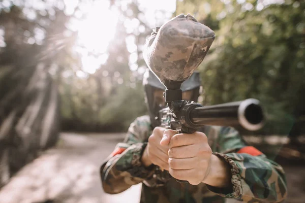 Selective focus of male paintball player in goggle mask and camouflage aiming by paintball gun outdoors — Stock Photo