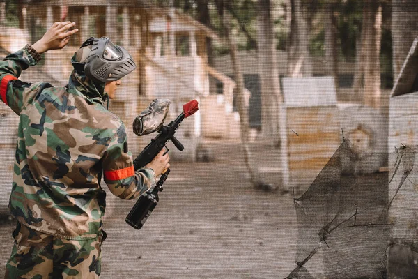 Vista trasera del jugador de paintball masculino en máscara de gafas y camuflaje con pistola de paintball de pie cerca de la red al aire libre - foto de stock