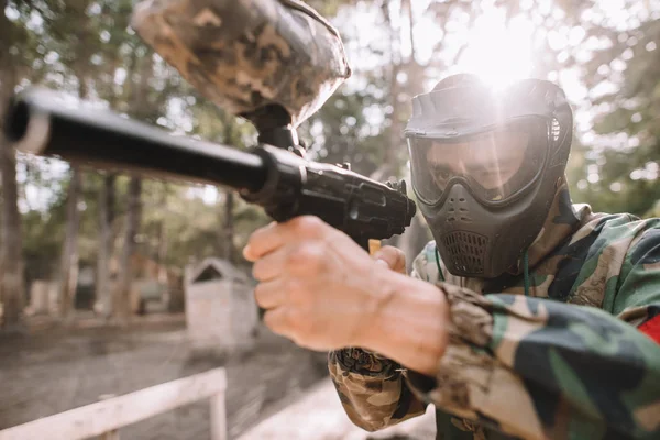 Focused male paintball player in goggle mask and camouflage aiming by paintball gun outdoors — Stock Photo