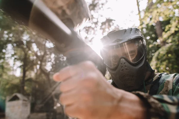 Selective focus of male paintball player in goggle mask and camouflage aiming by paintball gun outdoors — Stock Photo