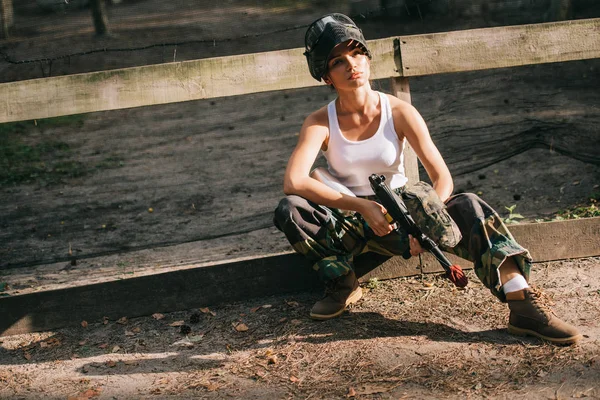 Joven mujer paintballer en singlet blanco y máscara de gafas que sostiene la pistola de paintball al aire libre - foto de stock