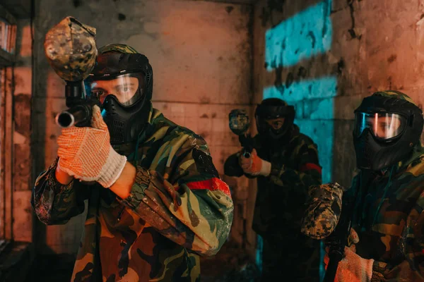 Paintball team in uniform and protective masks playing paintball with marker guns in abandoned building — Stock Photo