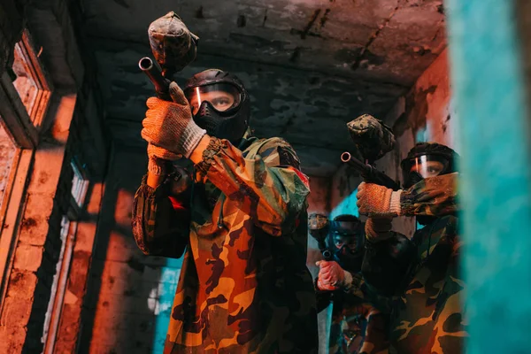 Low angle view of paintball team in uniform and protective masks aiming by paintball guns in abandoned building — Stock Photo