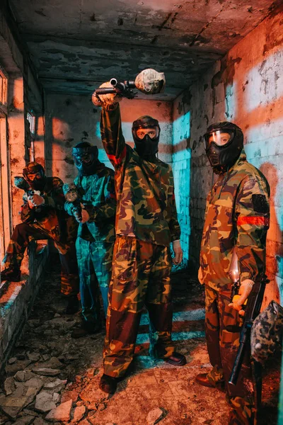 Чоловічий гравець в пейнтбол, спрямований на пістолет на камеру, в той час як його команда в пейнтболі в уніформі і захисних масках, що стоять поруч в покинутій будівлі — Stock Photo