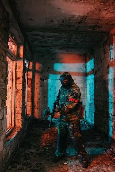 Jugador de paintball masculino en máscara de gafas y pistola de paintball de carga de camuflaje en edificio abandonado - foto de stock