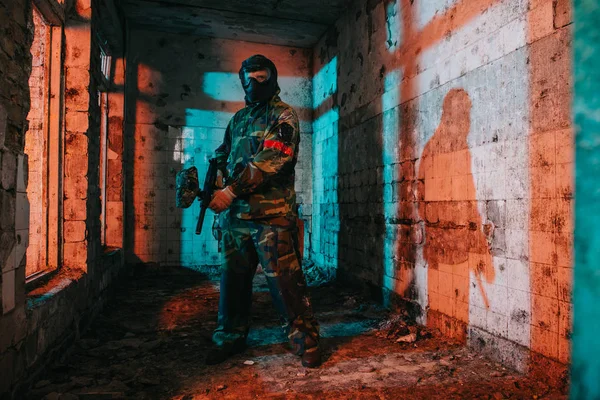 Joueur de paintball masculin confiant dans un masque à lunettes et un camouflage tenant un pistolet de paintball dans un bâtiment abandonné — Photo de stock