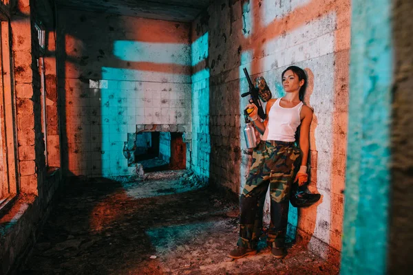 Jeune peintre féminine en camouflage et singulet blanc tenant pistolet de paintball et masque de masque dans un bâtiment abandonné — Photo de stock