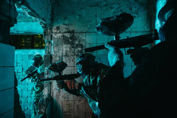 Paintball players in uniform and protective masks playing paintball with marker guns in abandoned building — Stock Photo